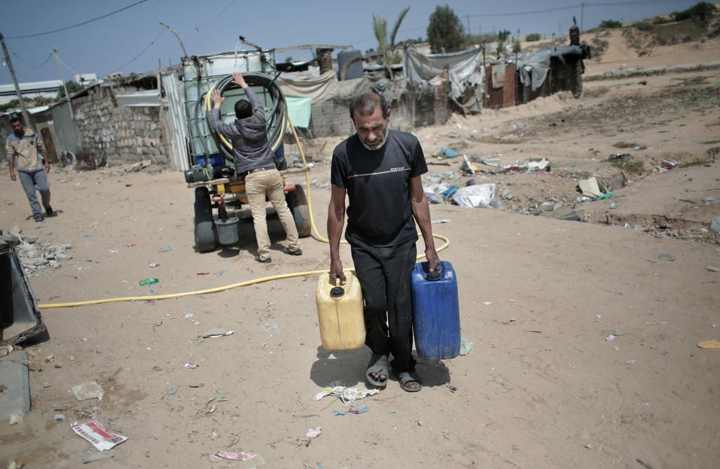 A Palestinian man carries plastic gallons he filled with drinking water from a vendor, background, in Khan Younis refugee camp, southern Gaza Strip. (AP Photo/Khalil Hamra)