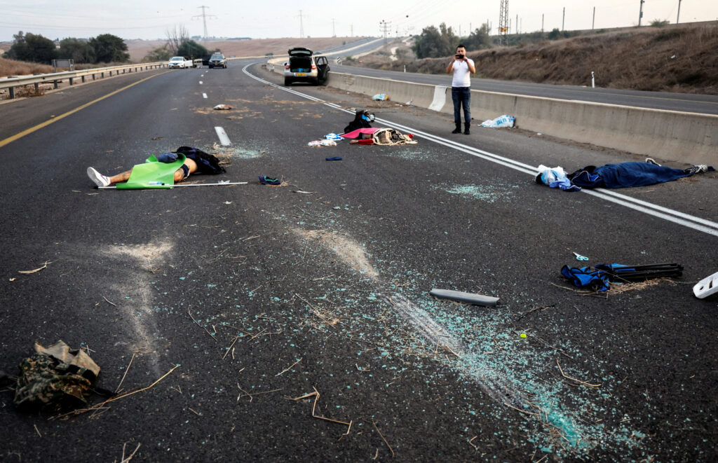 A man takes a photo of murder victims strewn across a road following a mass-infiltration by Hamas gunmen from the Gaza Strip, in the Sderot area, southern Israel, October 7, 2023 (Image: Reuters/Ammar Awad/ AAP)