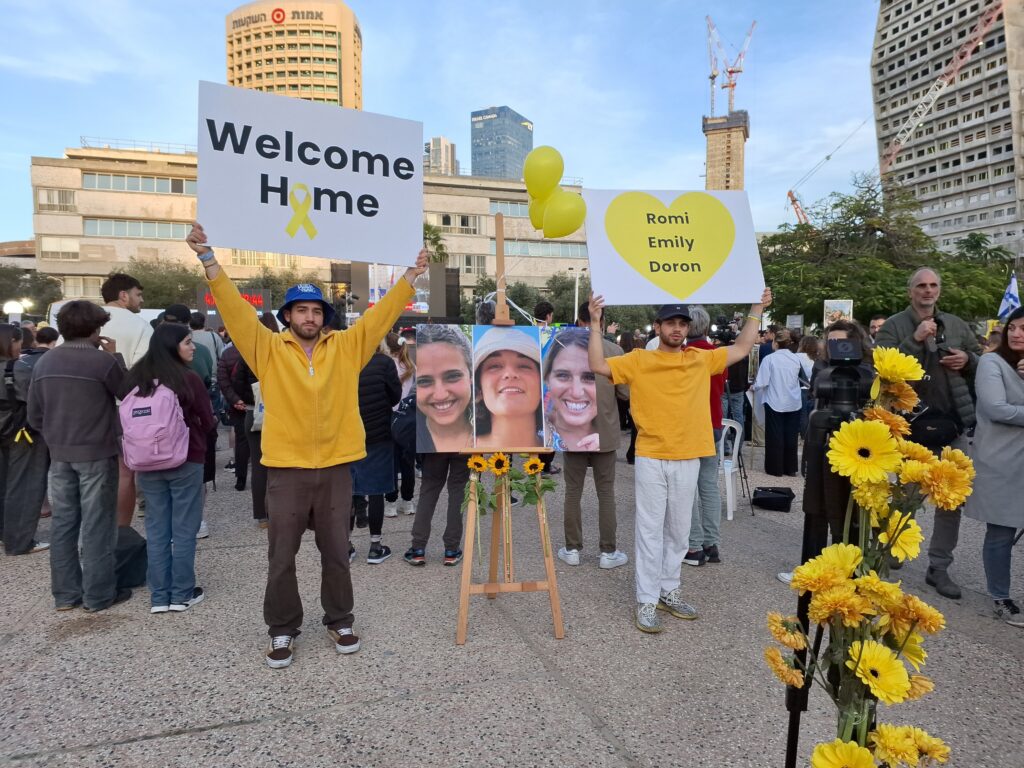 Israelis at Rabin Square holding welcome home signs next to photos of Romi Gonen, Emily Damari and Doron Steinbrecher, Tel Aviv, 19 January 2025 (Image: Vered Barequet/ Shutterstock)