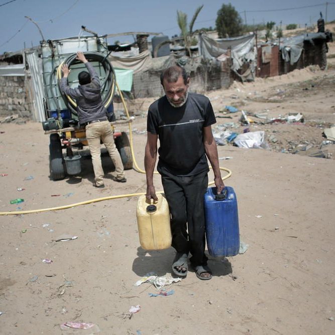 A Palestinian man carries plastic gallons he filled with drinking water from a vendor, background, in Khan Younis refugee camp, southern Gaza Strip. (AP Photo/Khalil Hamra)