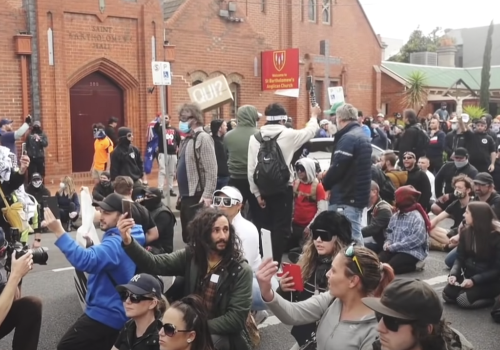 Protesters at a Melbourne anti-lockdown protest hold a poster with the French word “Qui” on it. “Qui” is a codeword used by white supremacists to imply Jews (Image: screenshot)