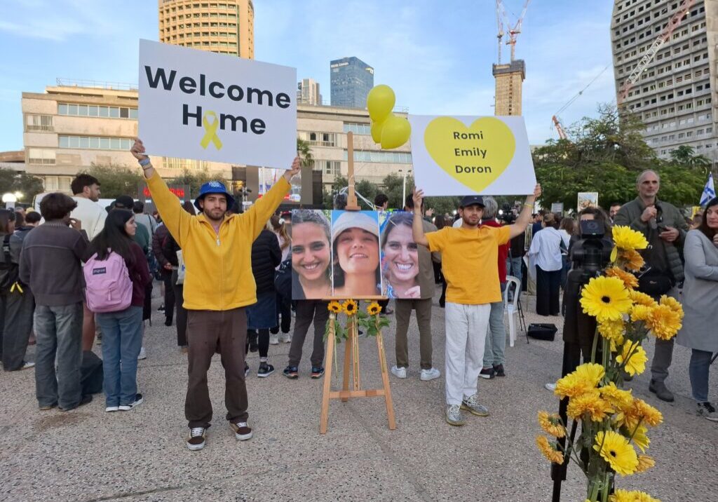 Israelis at Rabin Square holding welcome home signs next to photos of Romi Gonen, Emily Damari and Doron Steinbrecher, Tel Aviv, 19 January 2025 (Image: Vered Barequet/ Shutterstock)