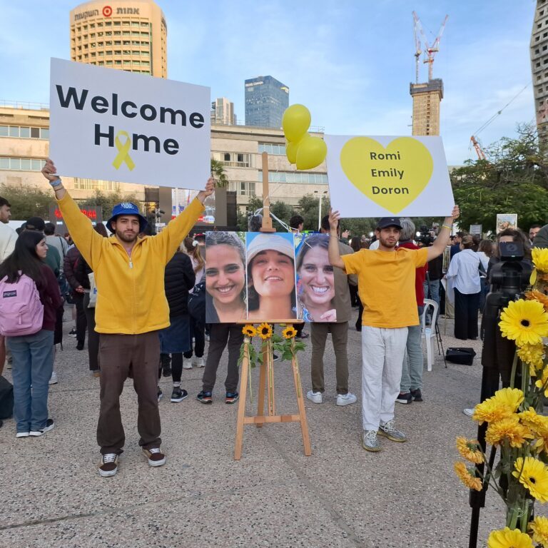 Israelis at Rabin Square holding welcome home signs next to photos of Romi Gonen, Emily Damari and Doron Steinbrecher, Tel Aviv, 19 January 2025 (Image: Vered Barequet/ Shutterstock)